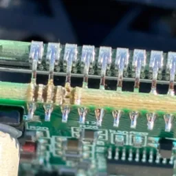 Zbliżenie na uszkodzone połączenia lutowane na zielonej płytce drukowanej w urządzeniu elektronicznym, widoczne ślady przegrzania i stopionego spoiwa.