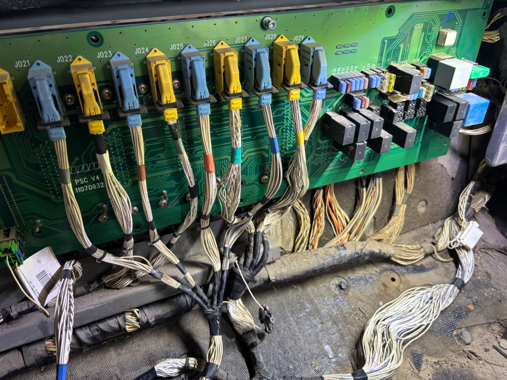 Zielona płyta obwodu drukowanego z rzędami kolorowych złączy i wiązkami kabli, widoczne liczne przekaźniki i bezpieczniki, całość w zakurzonym otoczeniu.
