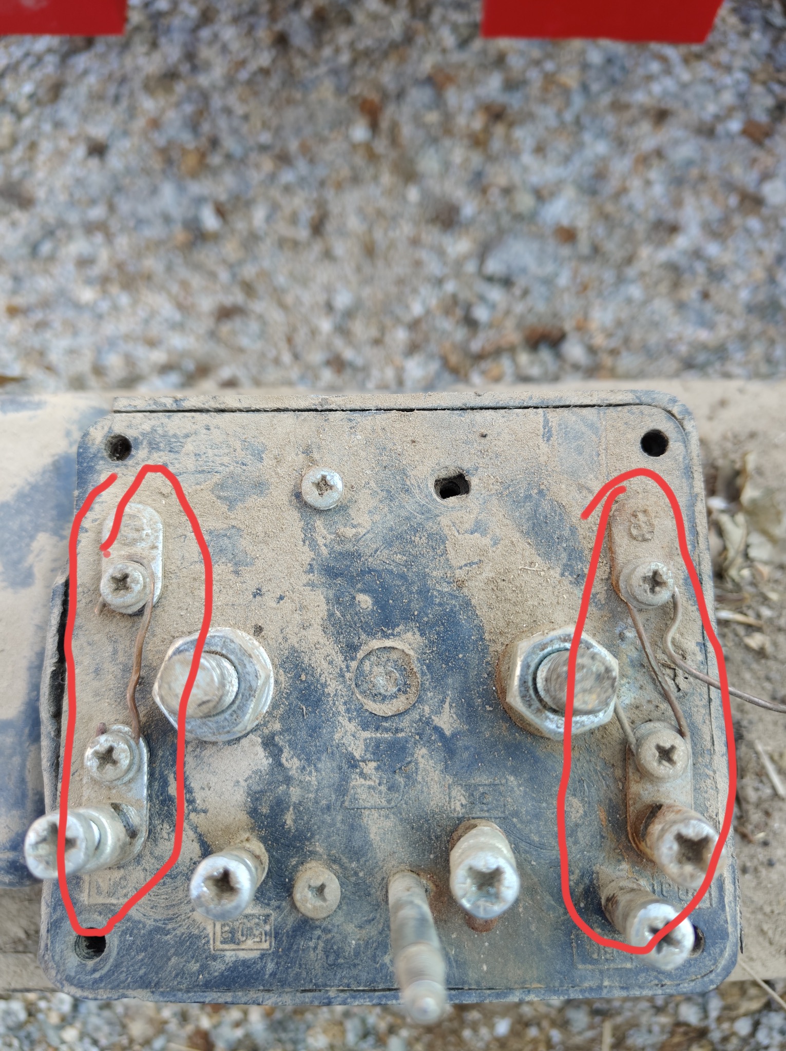 Brudny, zakurzony moduł elektryczny pojazdu z widocznymi śrubami, przewodami i elementami złącznymi, sfotografowany z góry na tle żwirowej powierzchni.