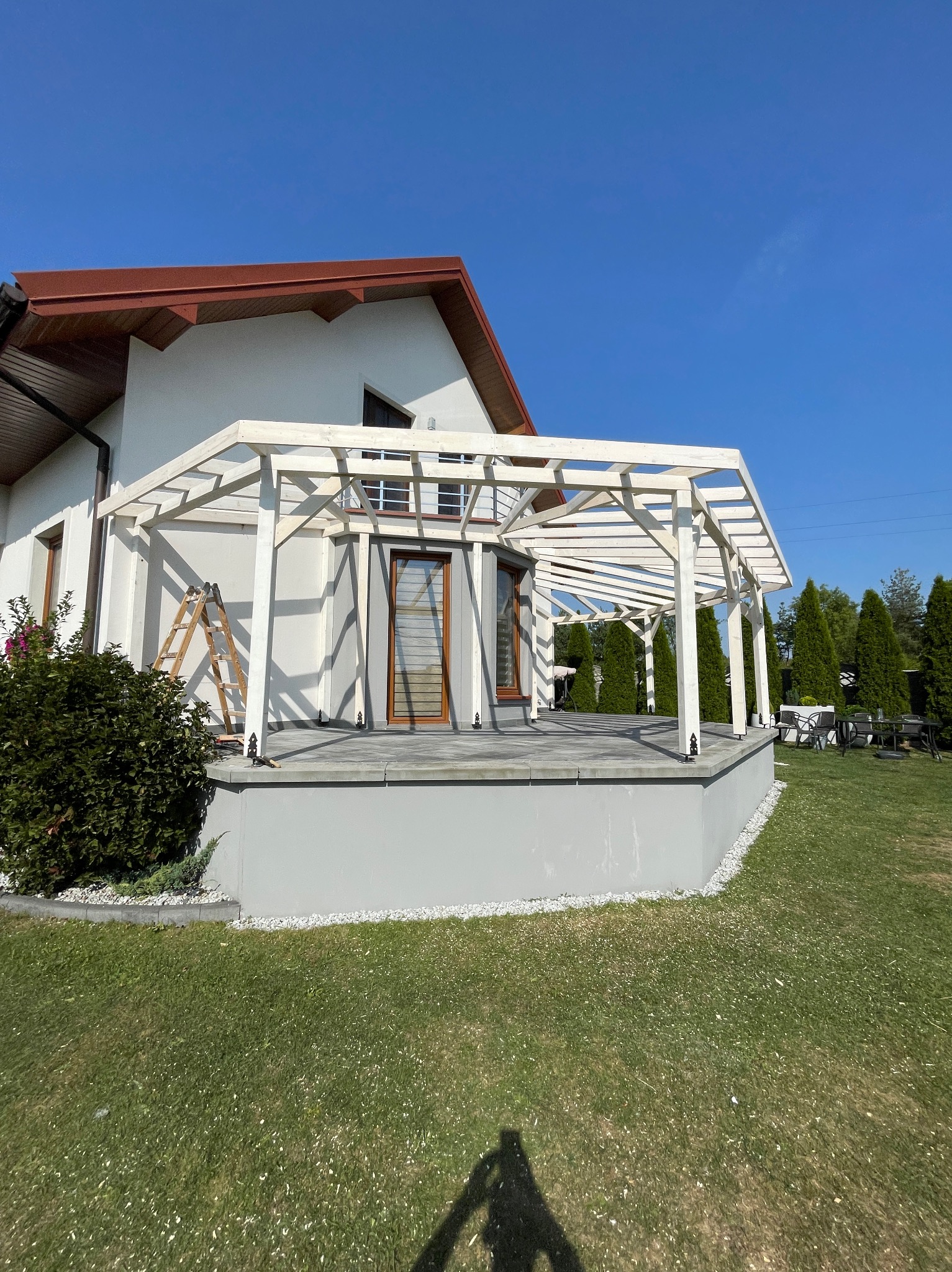 Biała, drewniana konstrukcja zadaszenia tarasu w trakcie budowy, oparta na szarym podeście, drabina oparta o ścianę domu, błękitne niebo w tle.