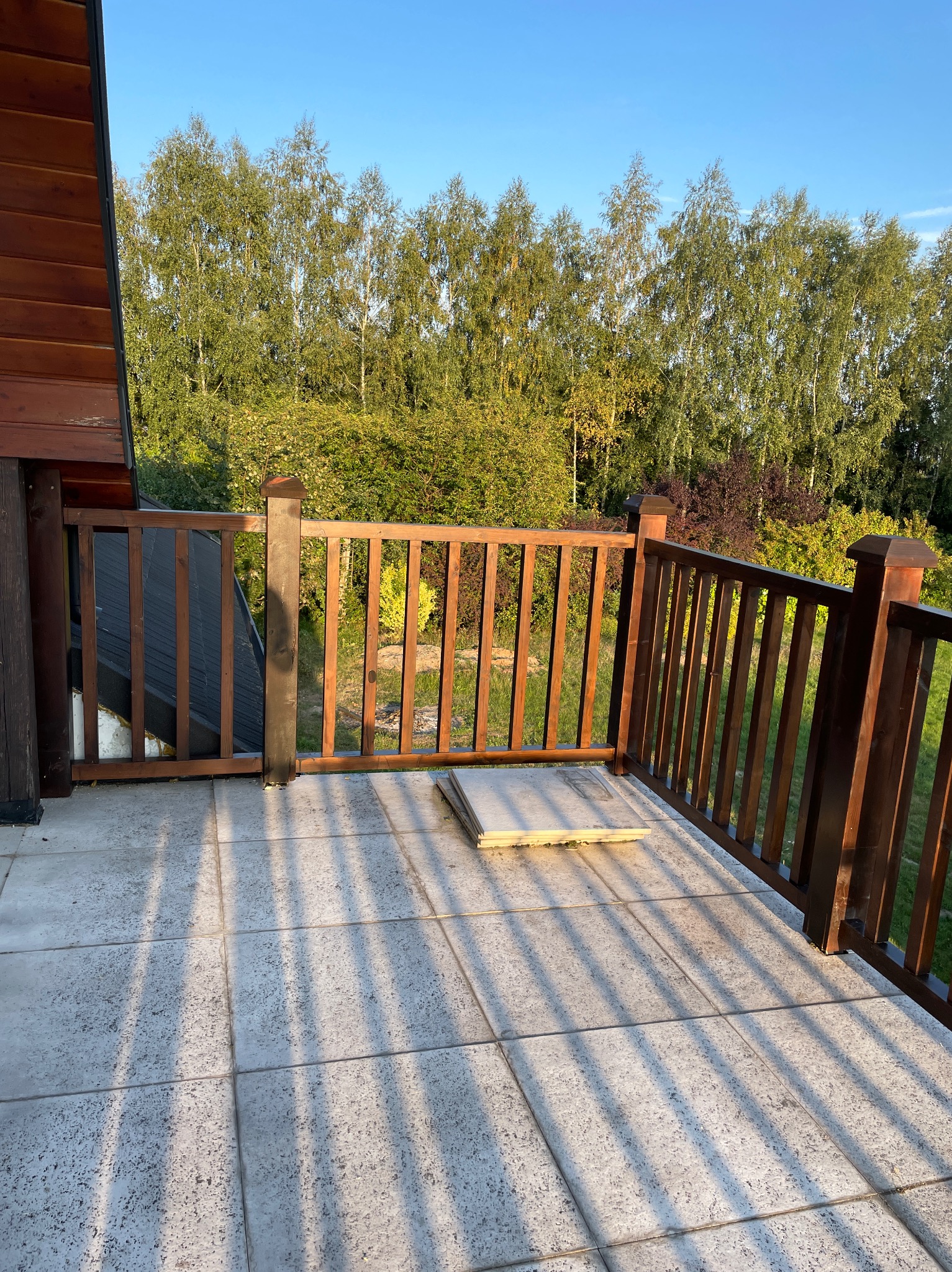 Barierka z ciemnego drewna na tarasie z jasnych płytek, w tle drzewa i niebieskie niebo.