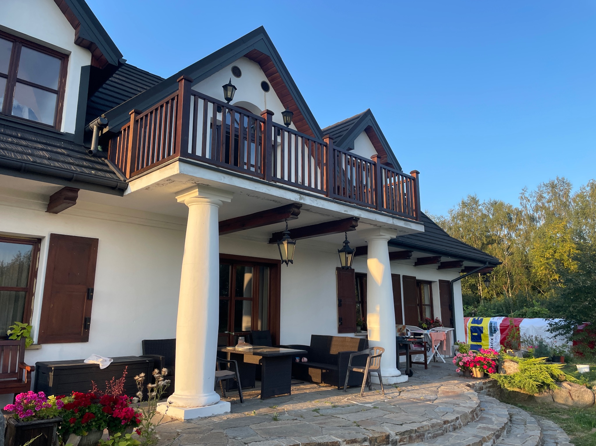 Dwukondygnacyjny dom z drewnianą balustradą na balkonie, kolumnami podtrzymującymi, kamiennym tarasem i meblami ogrodowymi.