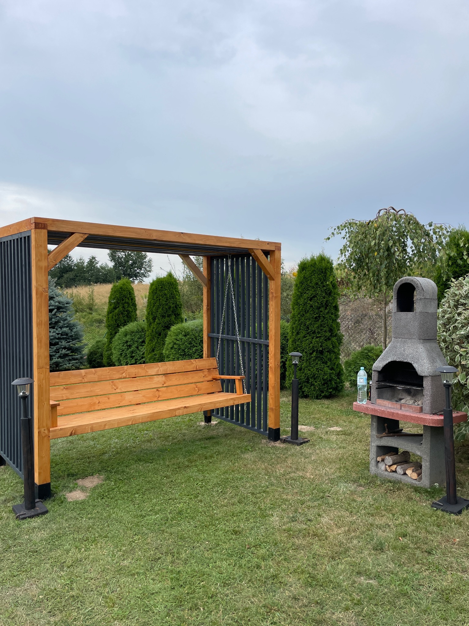 Drewniana huśtawka ogrodowa z zadaszeniem i elementami metalowymi, obok murowany grill z zapasem drewna, na trawniku w ogrodzie.