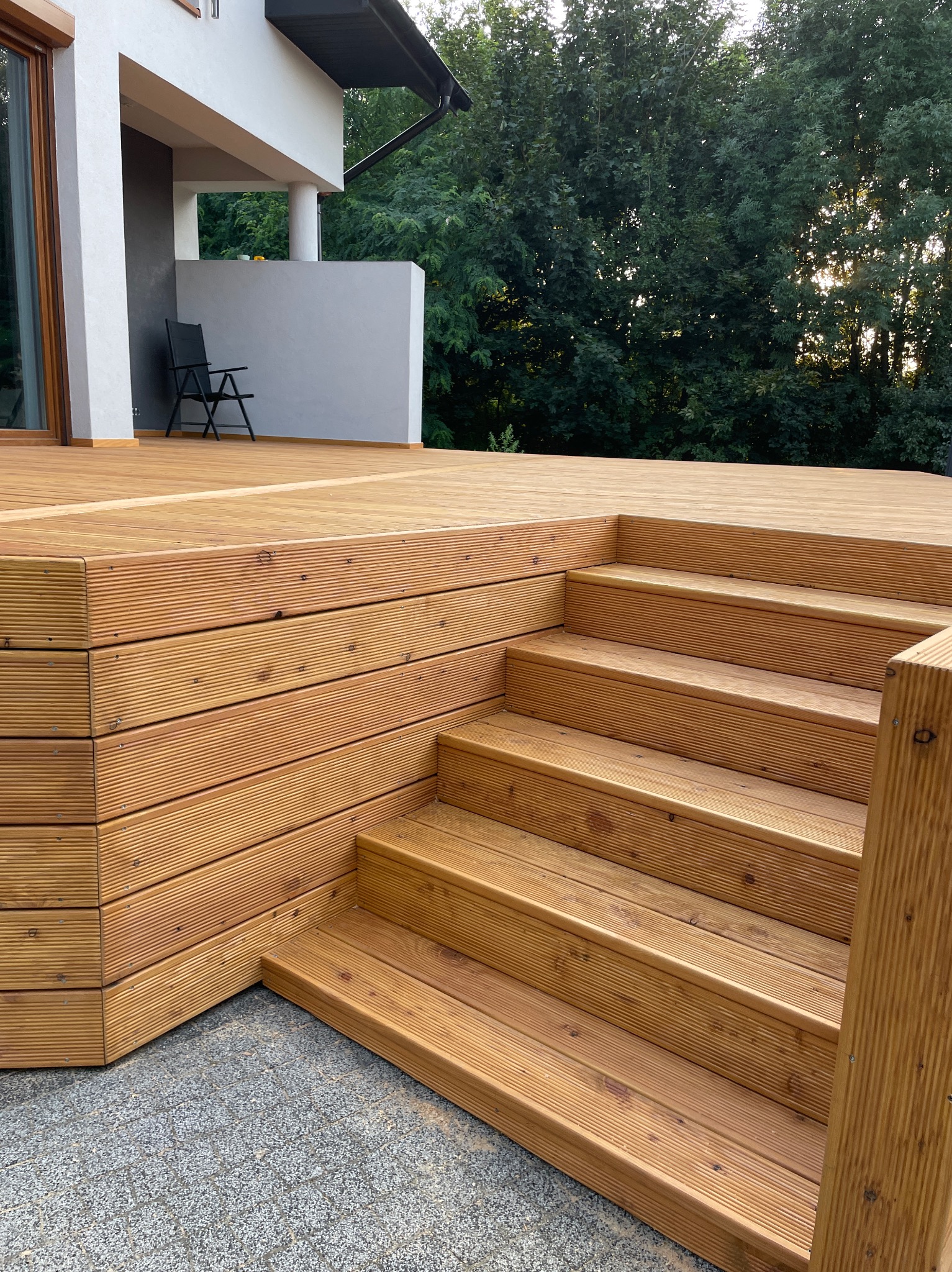 Nowoczesny taras z drewna z niskimi schodami, widok z dołu na jasne deski, w tle dom z balkonem i pojedyncze krzesło.