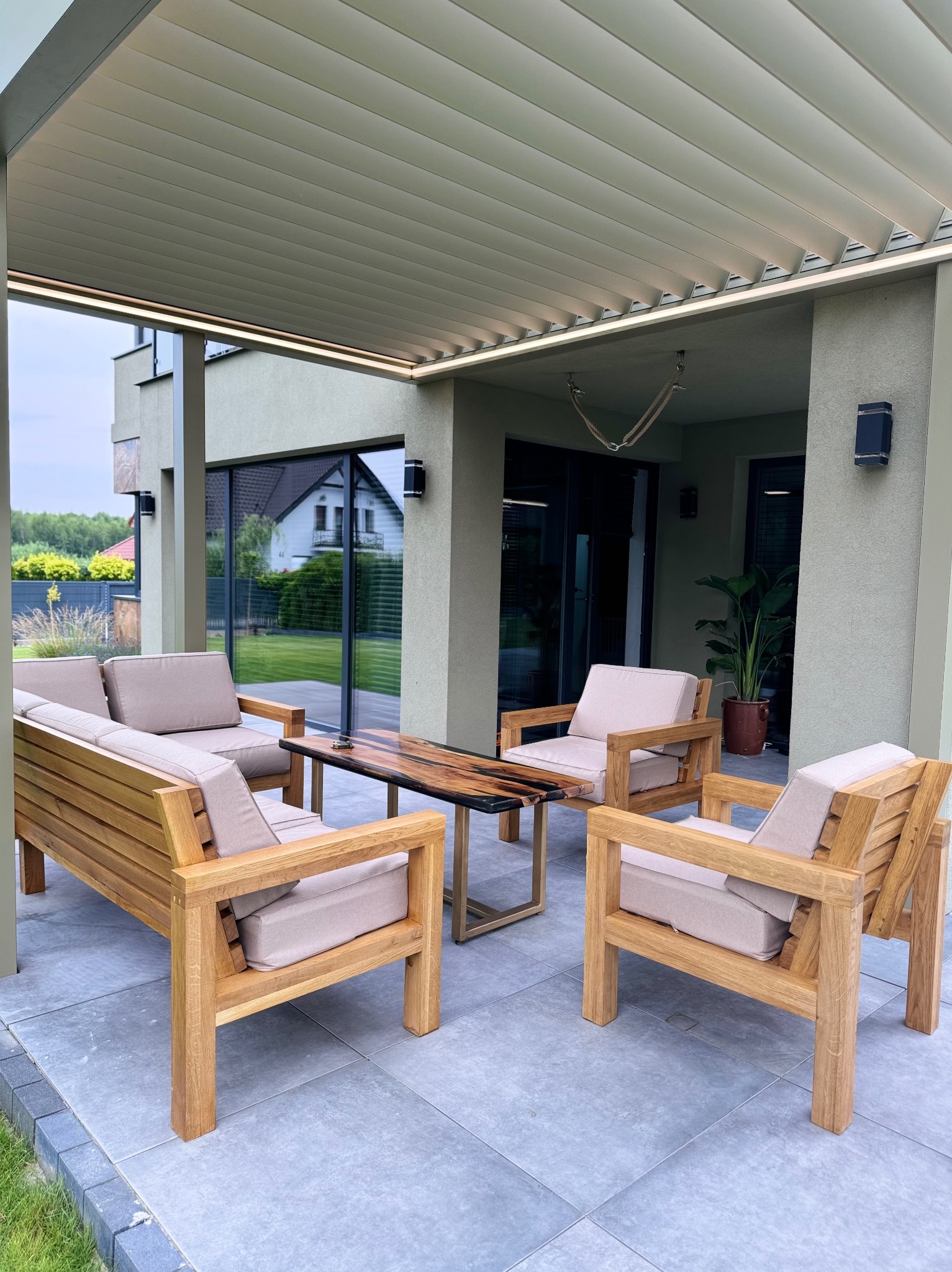 Zadaszony taras z drewnianymi meblami ogrodowymi: sofa, dwa fotele i stolik kawowy z żywicą epoksydową, szara podłoga z płytek, nowoczesny dom w tle.