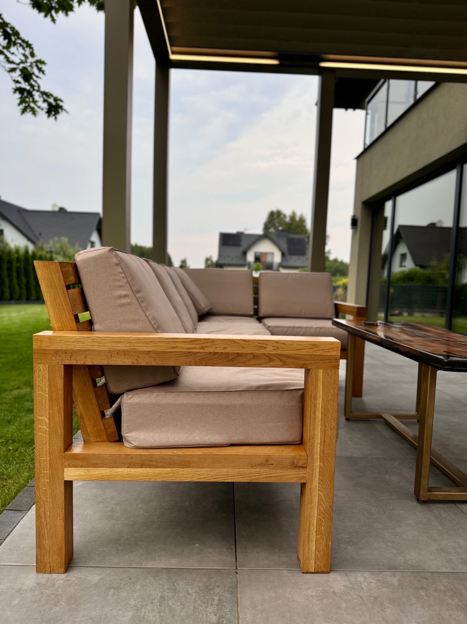Drewniany narożnik z beżowymi poduchami na zadaszonym tarasie, obok stolik kawowy z blatem z żywicy epoksydowej, w tle dom z dużymi oknami.