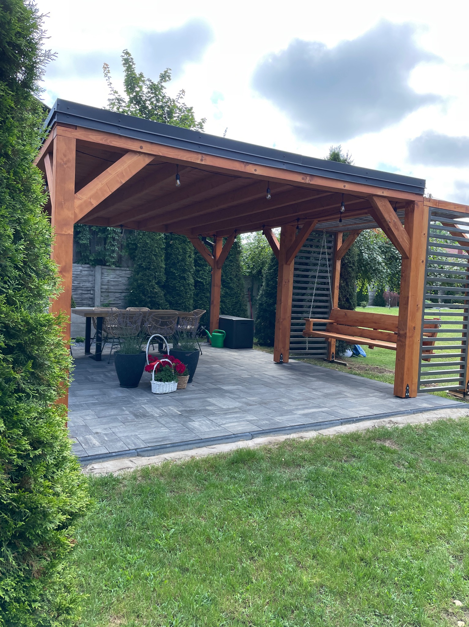 Zadaszona konstrukcja z drewna, z huśtawką, stołem i krzesłami, na wyłożonym kostką brukową patio, otoczona zielenią.