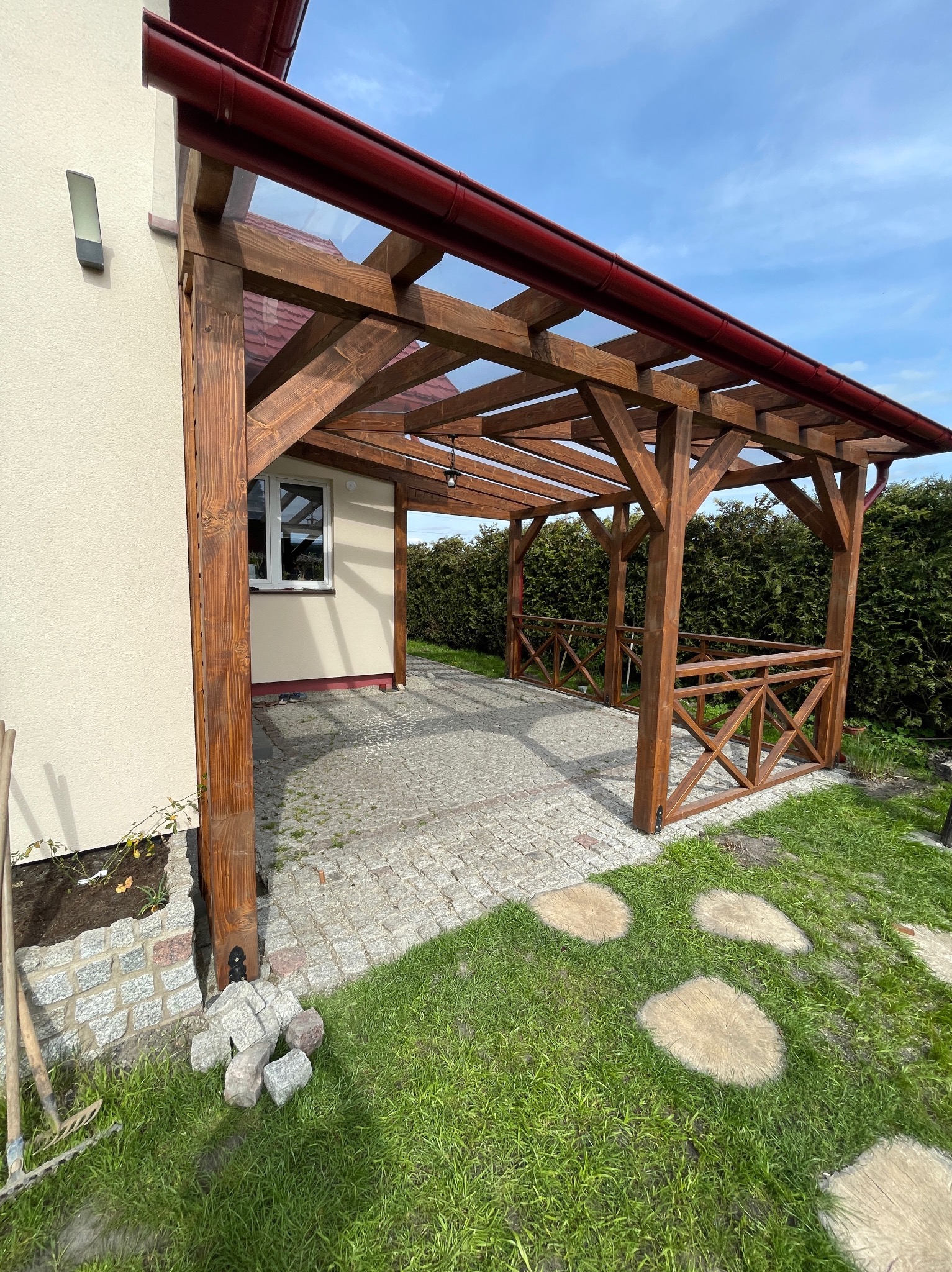 Drewniana konstrukcja z przezroczystym zadaszeniem przylegająca do budynku, z niską balustradą, wyłożona kostką brukową, z elementami krajobrazu: trawnik, ścieżka z pniaków, kamienie i narzędzia...