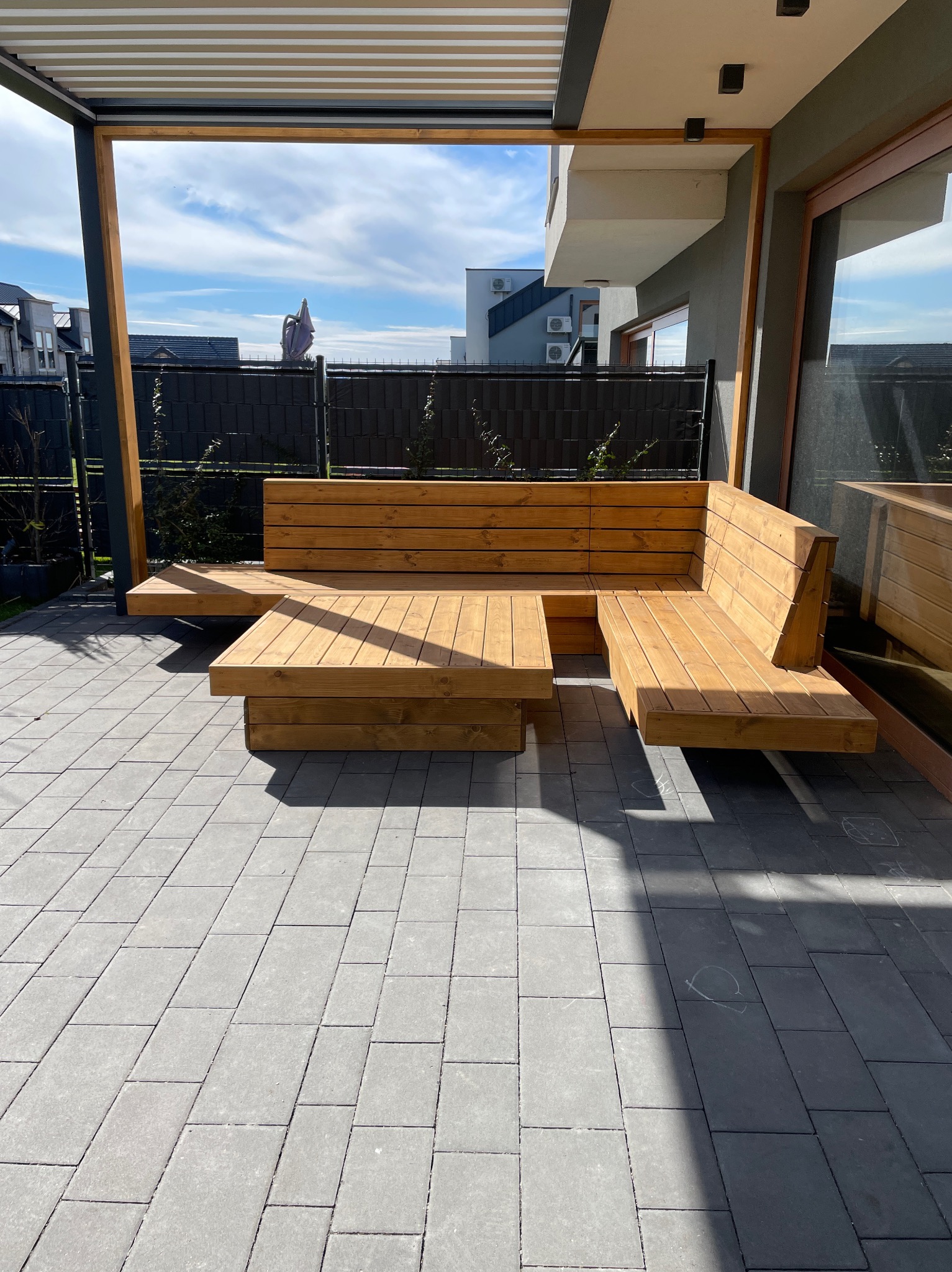 Drewniany narożnik z niskim stolikiem pod pergolą na patio z szarej kostki brukowej, widok na nowoczesny dom i ogrodzenie.