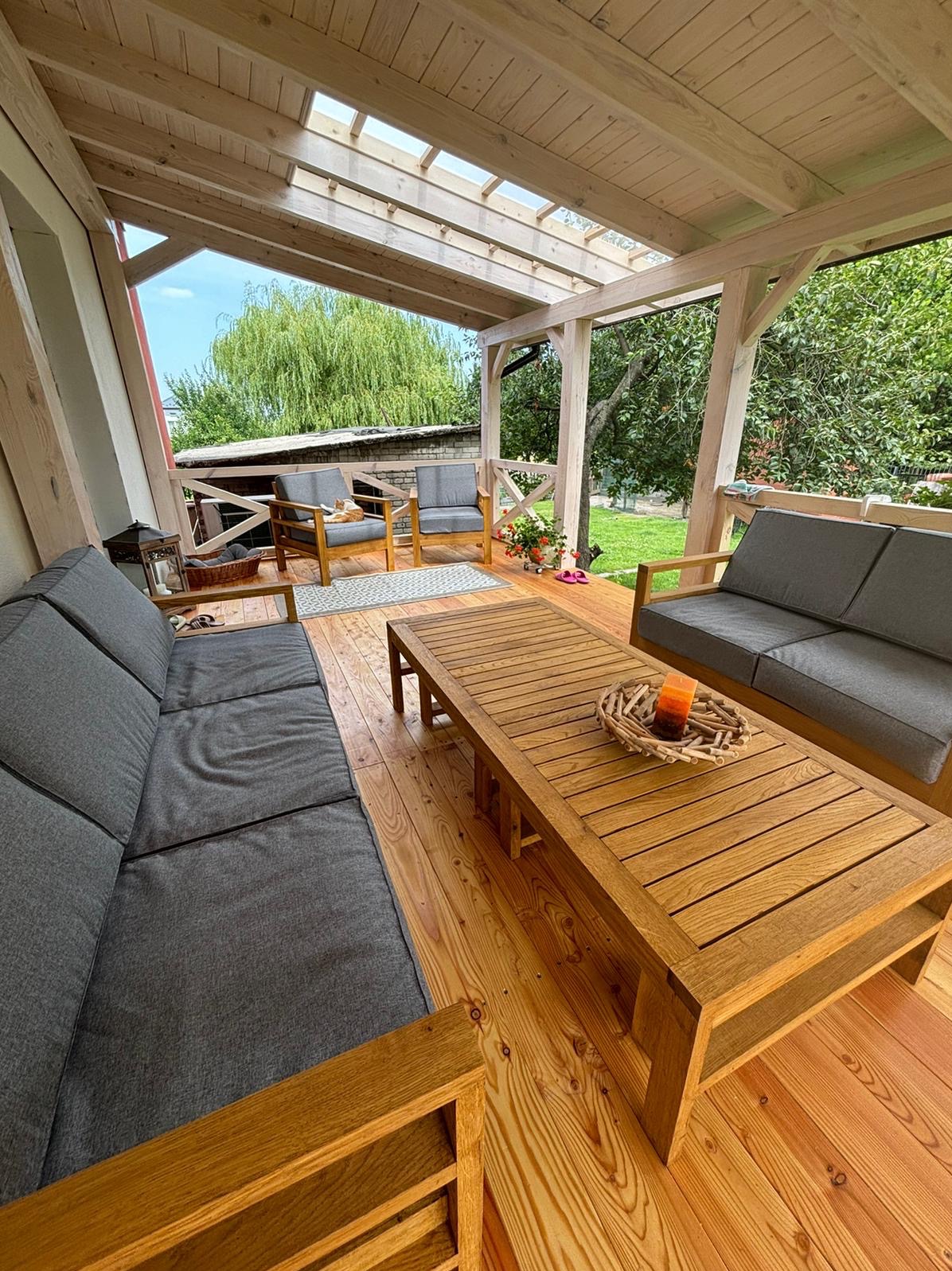 Zadaszony taras z drewnianą podłogą i meblami ogrodowymi: szarą sofą, fotelami i stolikiem kawowym z dekoracją ze świecą. W tle widoczny ogród z drzewem płaczącą wierzbą.
