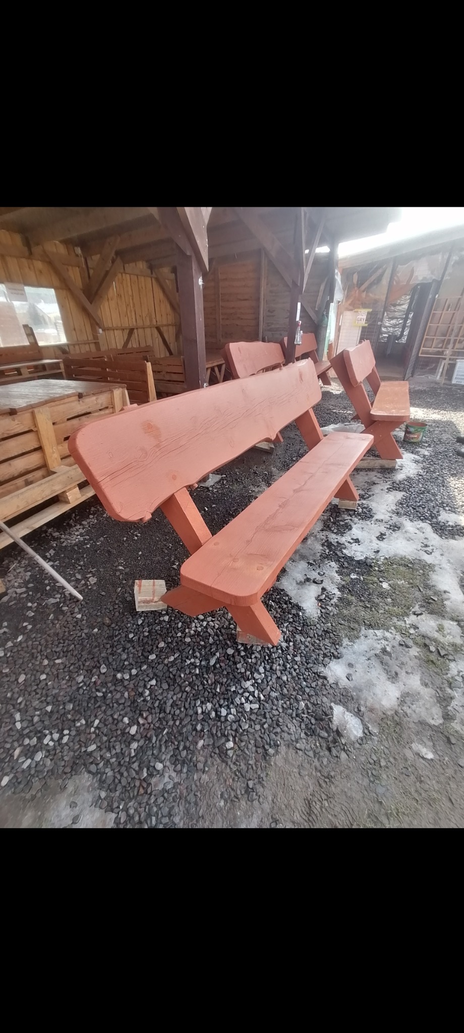 Rustykalne, pomalowane na pomarańczowo drewniane ławki z oparciami, ustawione na żwirowym podłożu z resztkami śniegu, pod drewnianą konstrukcją z widocznymi paletami w tle.