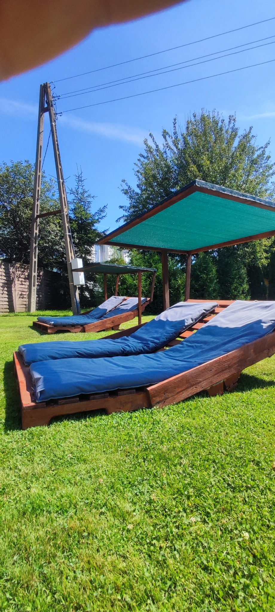 Trzy drewniane leżaki z niebieskimi materacami na trawniku, osłonięte zielonym zadaszeniem, w tle słup elektryczny.