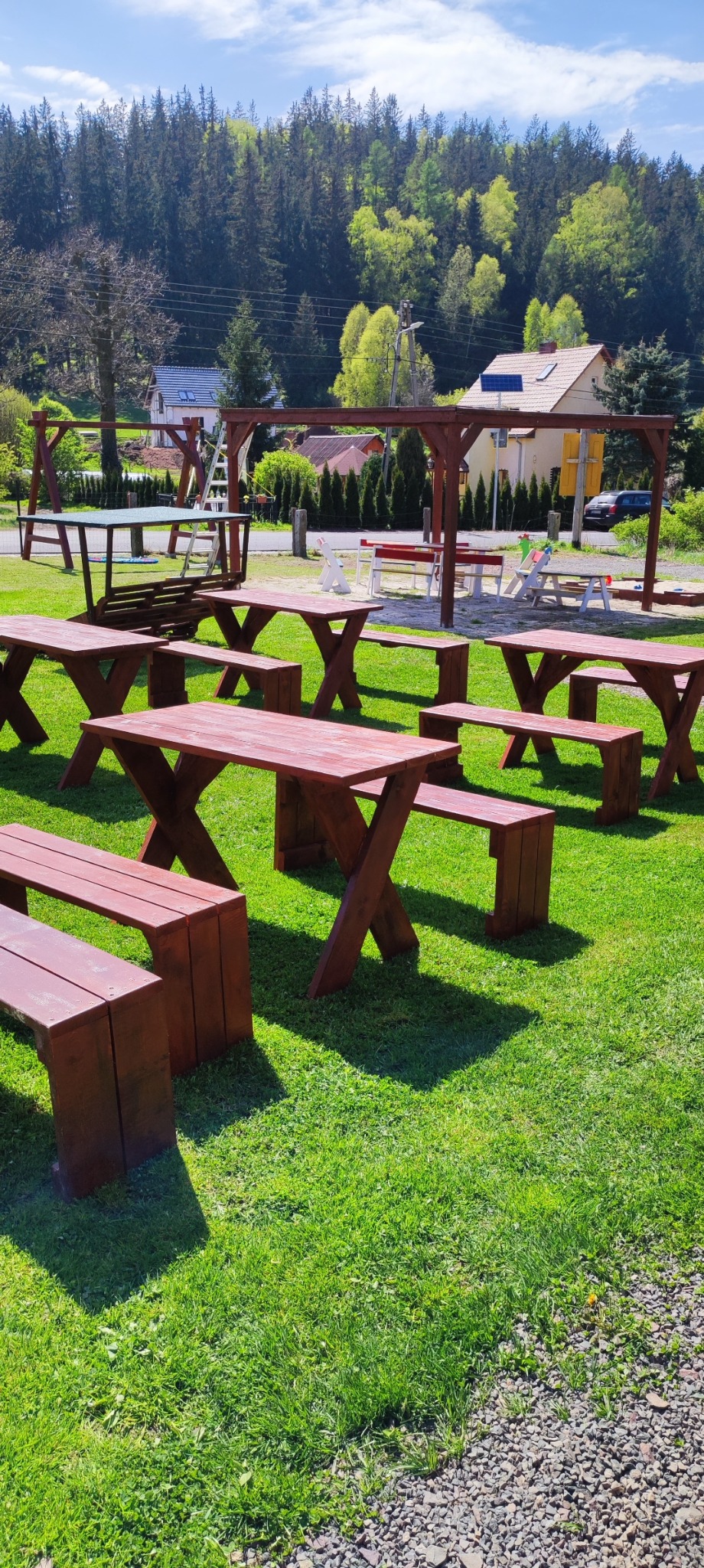 Zestaw drewnianych stołów i ławek na trawniku w Mieroszowie, z widoczną w tle konstrukcją z huśtawką i zadaszeniem, całość w słoneczny dzień.