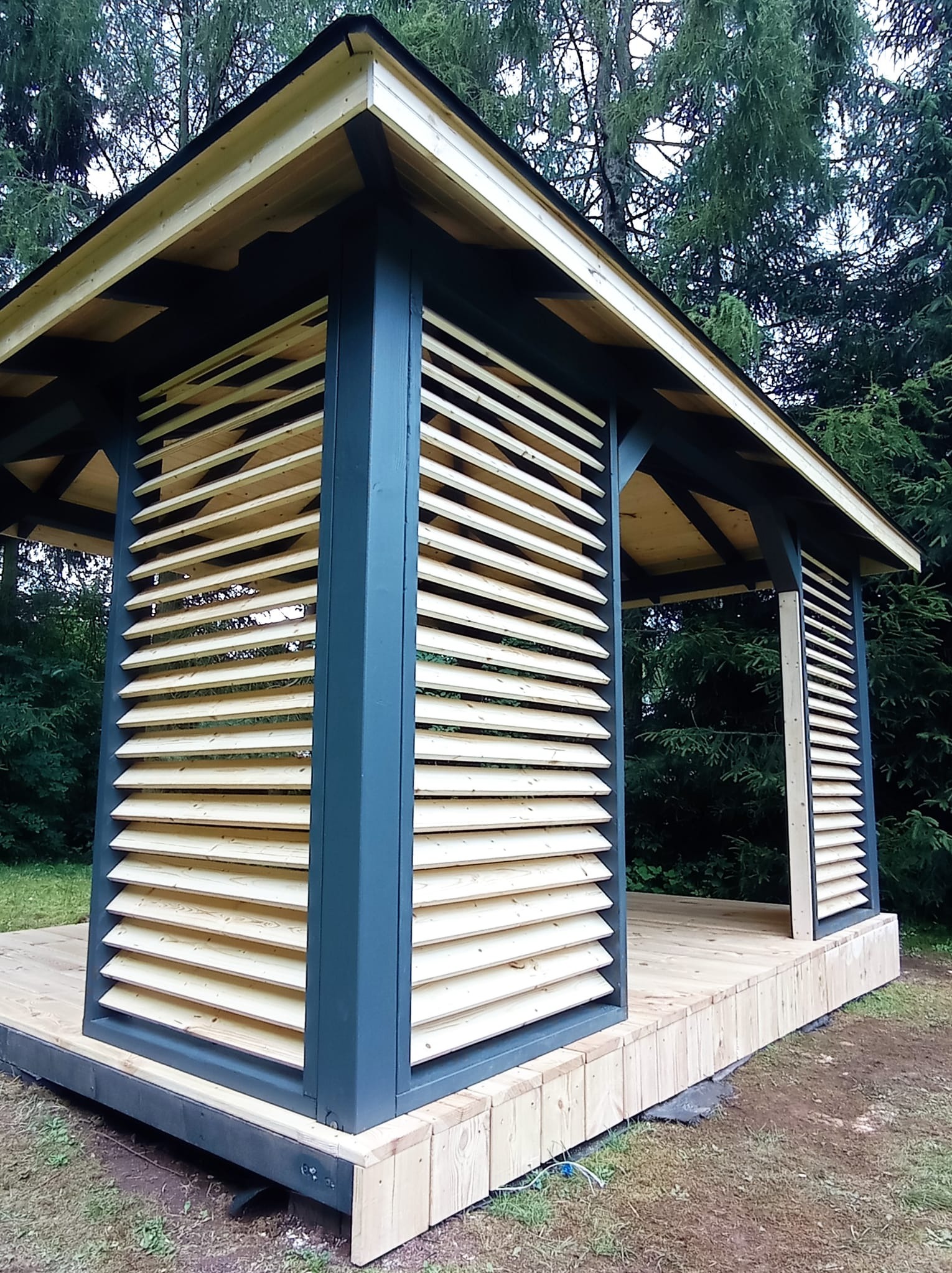 Drewniana altana z lamelami w kolorze naturalnego drewna i grafitowymi słupami, w tle iglaste drzewa. Widok z zewnątrz, w dziennym świetle.