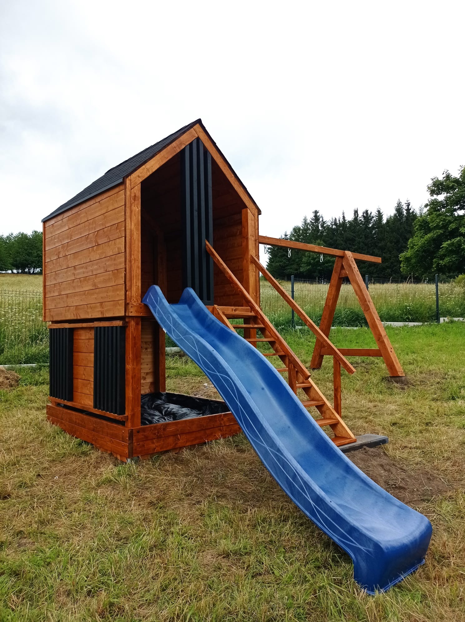 Drewniany plac zabaw z domkiem, zjeżdżalnią i huśtawką, w ogrodzie. Elementy pomalowane na brązowo i czarno. Zjeżdżalnia niebieska, z białym wzorem.