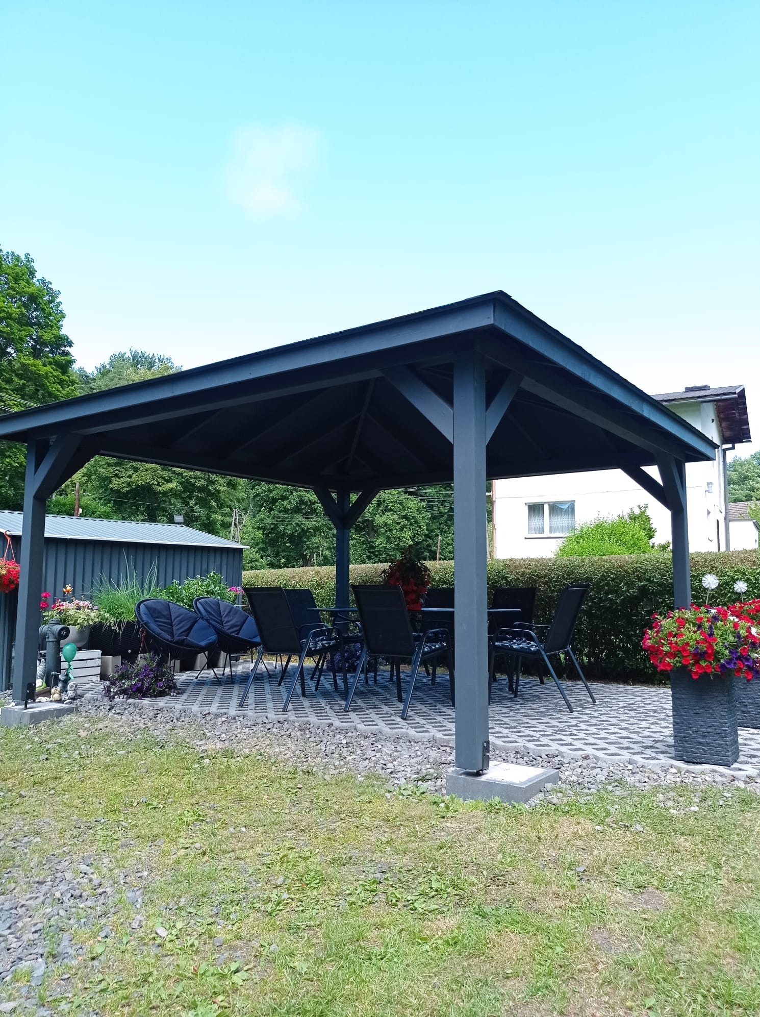 Szara altana ogrodowa z ciemnymi meblami ogrodowymi na kostce brukowej, otoczona zielenią. W tle widoczny budynek mieszkalny i krzewy.