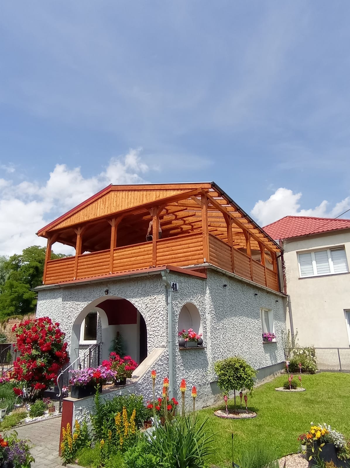 Drewniana konstrukcja z balustradą na dachu domu, widok z dołu. Konstrukcja z jasnego drewna, zadaszona, z widocznym numerem domu 41 i kwiatami w otoczeniu.