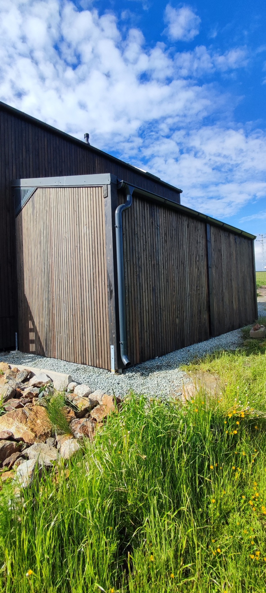 Nowoczesna, drewniana konstrukcja z pionowymi panelami i szarą rynną, otoczona zielenią i kamieniami. Błękitne niebo z chmurami w tle.
