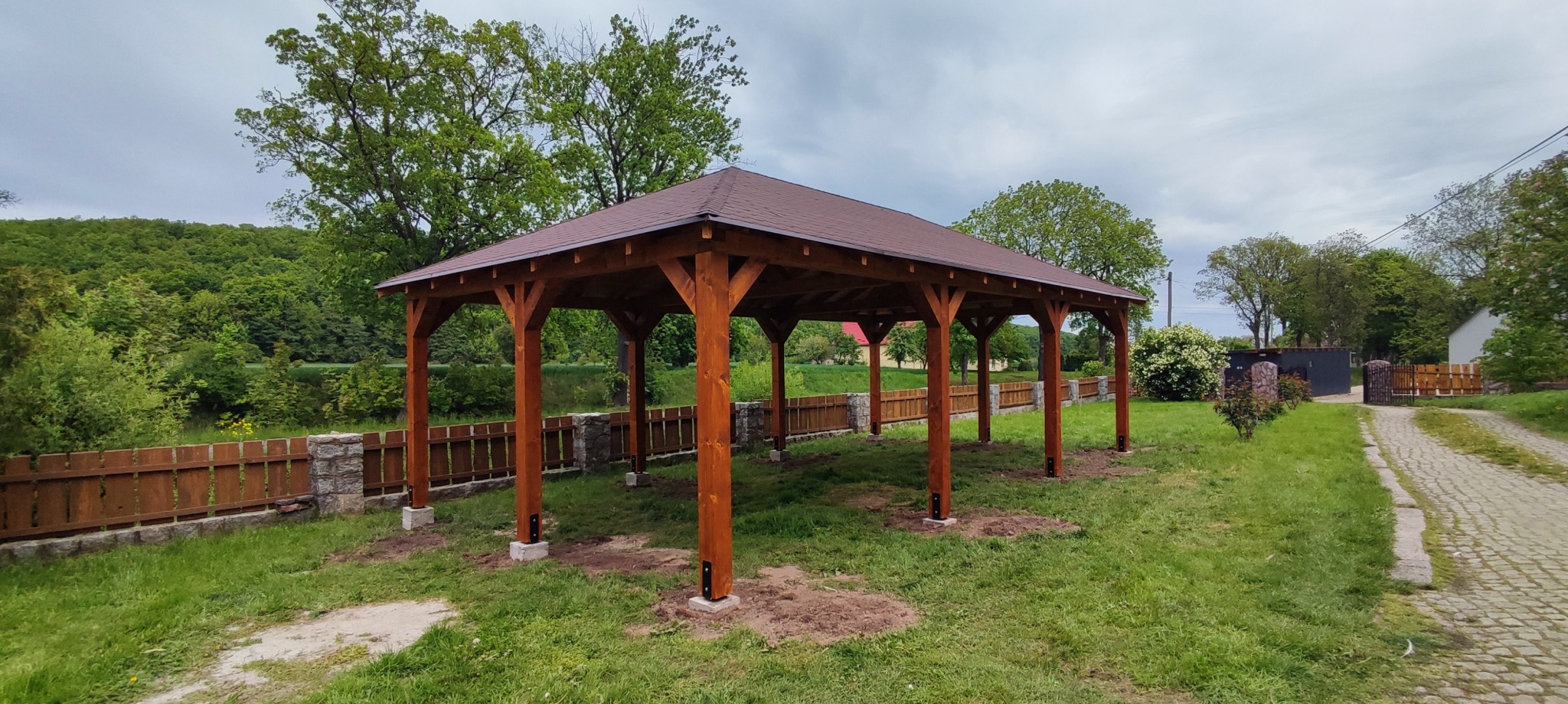 Brązowa, drewniana altana z dachem z gontu bitumicznego, na betonowych podstawach, otoczona zielenią i niskim drewnianym płotem z kamiennymi słupkami.