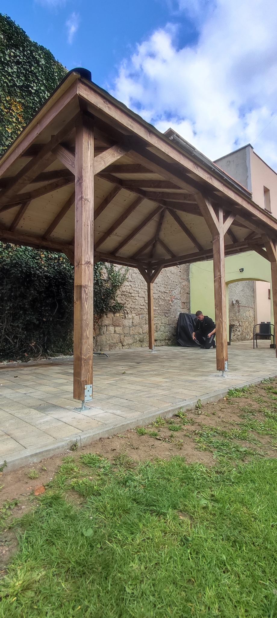 Ukończona, drewniana altana z ciemnego drewna na kostce brukowej. Widoczny mężczyzna w ciemnym ubraniu. Tło: mur z kamienia i budynek.