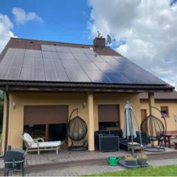 Dom jednorodzinny z brązowym dachem i panelami fotowoltaicznymi na dachu, widok z ogrodu, z tarasem z meblami ogrodowymi i dekoracjami.