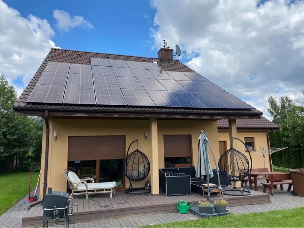 Dom jednorodzinny z brązowym dachem i panelami fotowoltaicznymi na dachu, widok z ogrodu, z tarasem z meblami ogrodowymi i dekoracjami.