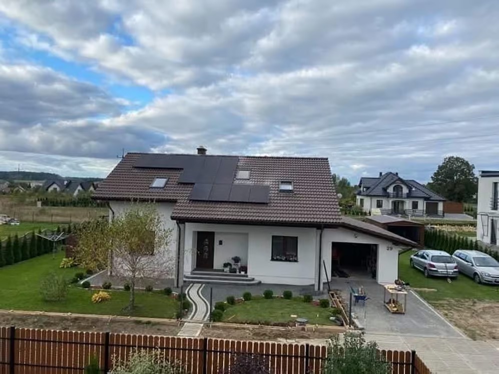 Dom jednorodzinny z brązowym dachem i panelami fotowoltaicznymi, widok z góry na uporządkowany ogród i podjazd z dwoma samochodami osobowymi.