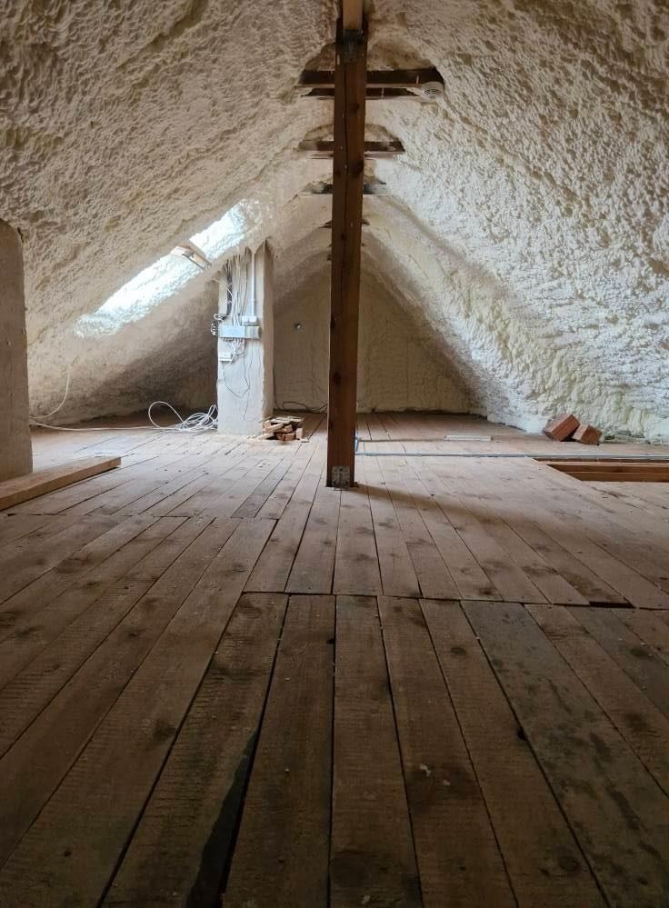 Poddasze z drewnianą podłogą i izolacją natryskową pianką PUR na skośnych ścianach i suficie, widoczne okno dachowe i pionowa belka konstrukcyjna.