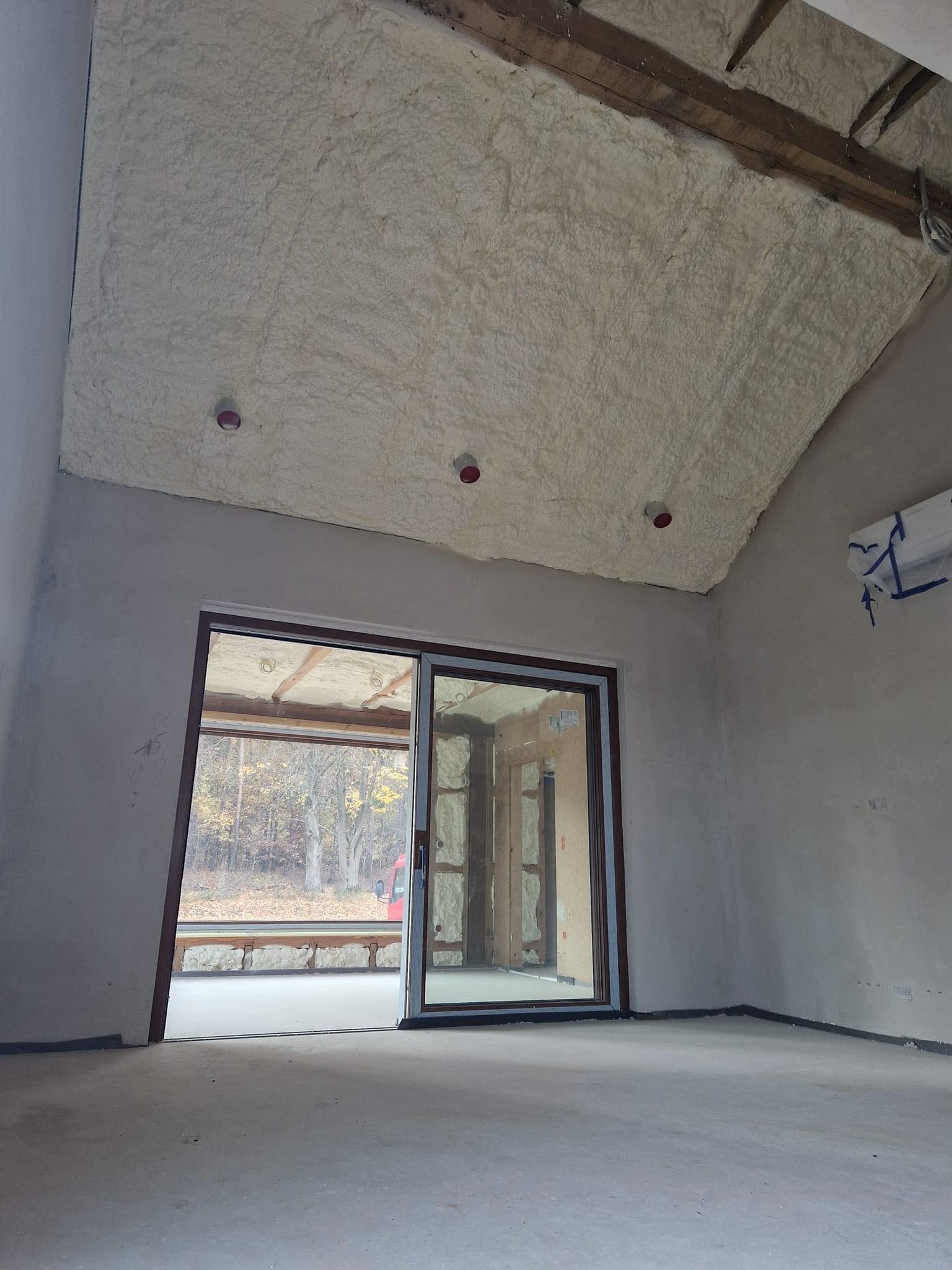 Poddasze domu w trakcie izolacji pianą PUR, widoczne drewniane belki stropowe, okno przesuwne z widokiem na jesienny krajobraz.