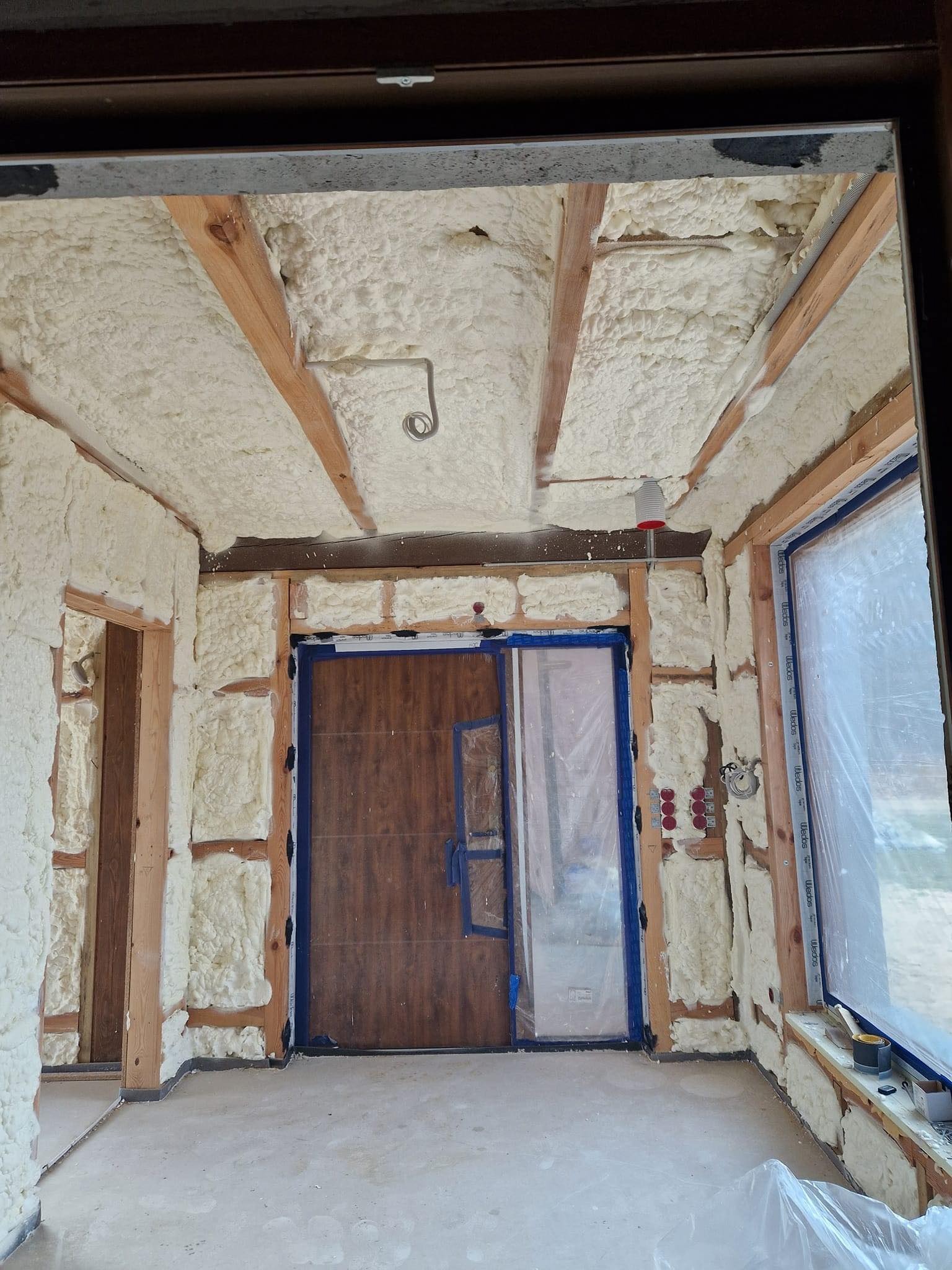 Piana izolacyjna wypełniająca przestrzeń między drewnianymi belkami w trakcie ocieplania poddasza, widoczne elementy instalacji elektrycznej i okna zabezpieczone folią.