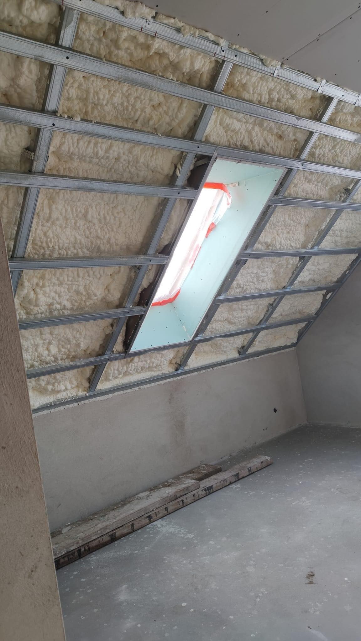 Poddasze w trakcie ocieplania pianą poliuretanową, widoczna metalowa konstrukcja sufitu podwieszanego i okno dachowe zabezpieczone folią.