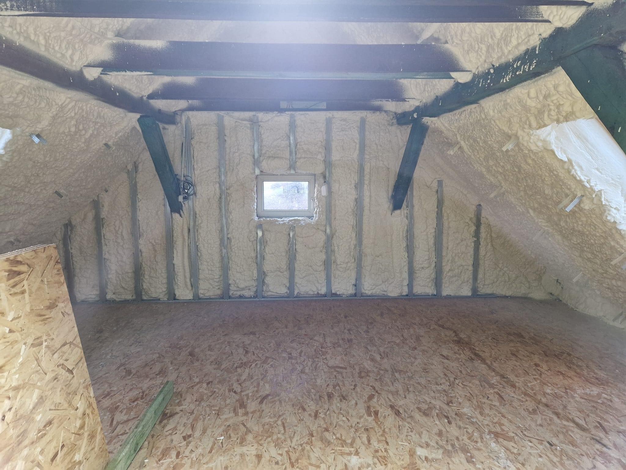 Poddasze z izolacją natryskową pianką poliuretanową na ścianach i skosach, widoczne metalowe profile i małe okno, podłoga wyłożona płytą OSB.