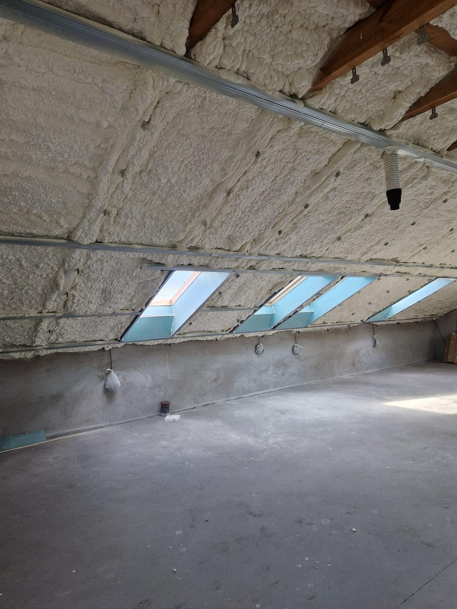 Poddasze z izolacją natryskową pianką poliuretanową, widoczne okna dachowe, surowe ściany i betonowa podłoga.