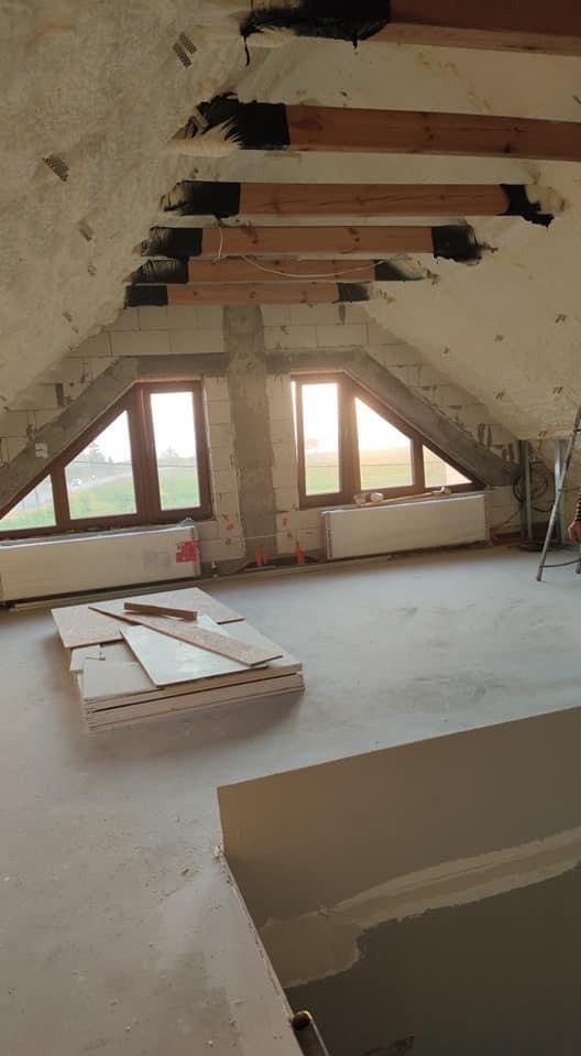 Poddasze w trakcie ocieplania i wykańczania: widoczne drewniane belki, izolacja piankowa, okna dachowe, grzejniki oraz stos płyt gipsowo-kartonowych.