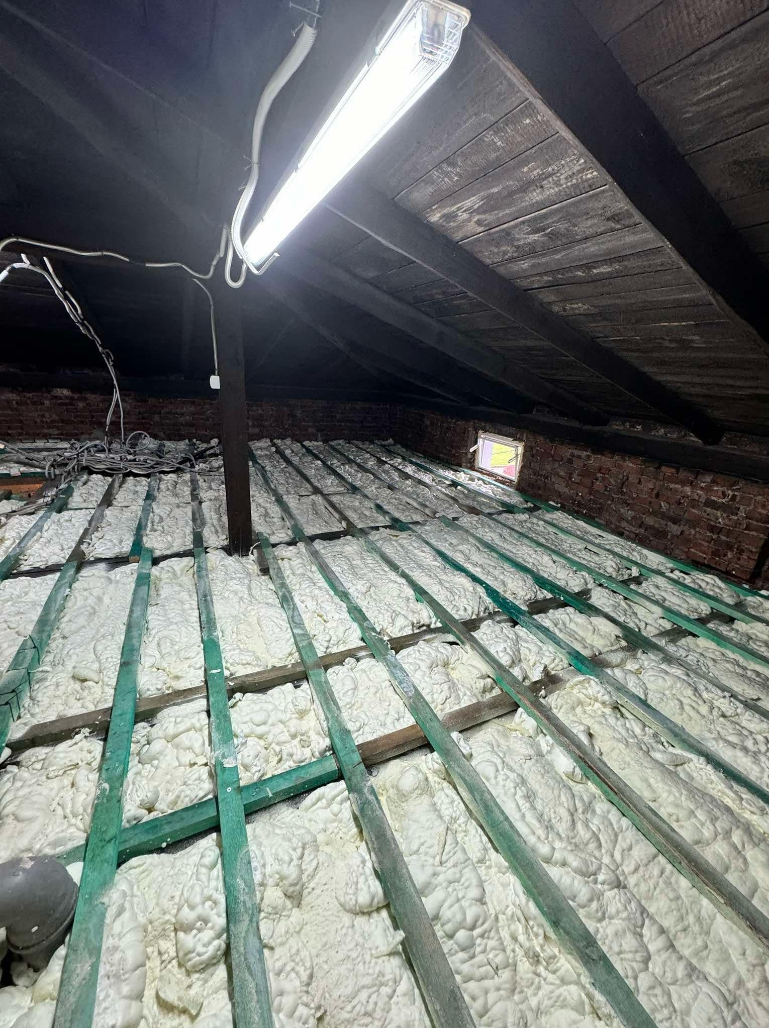 Izolacja poddasza pianą PUR między drewnianymi krokwiami, widoczne przewody elektryczne i lampa sufitowa