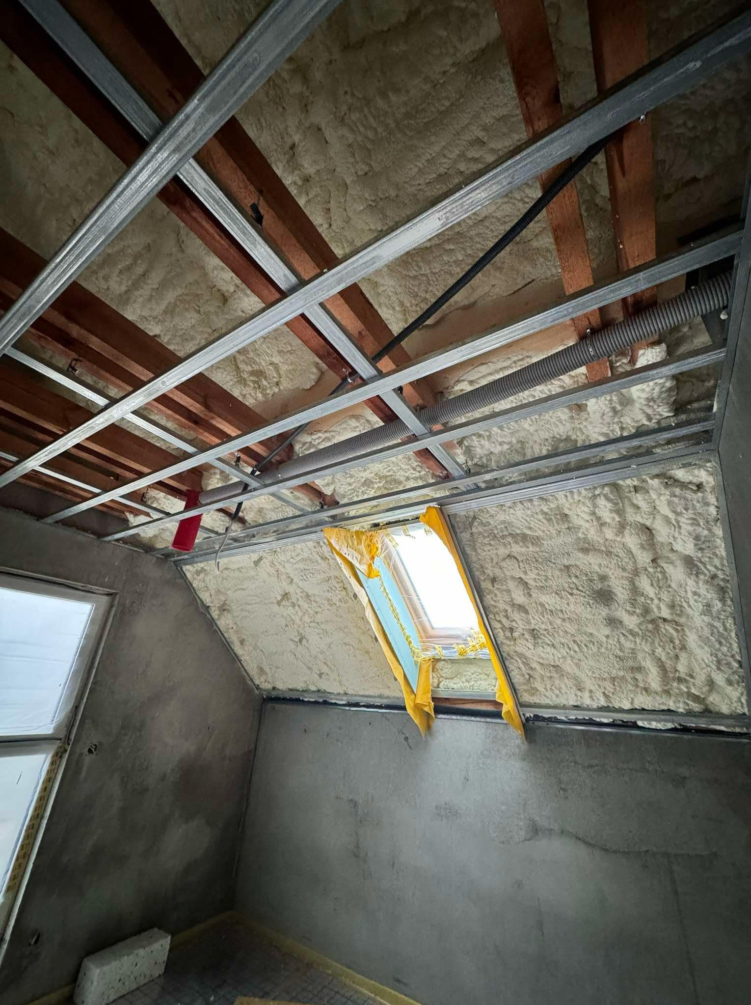 Poddasze w trakcie ocieplania pianką poliuretanową, widoczne metalowe profile konstrukcyjne sufitu, okno dachowe zabezpieczone folią, surowe ściany w tle.