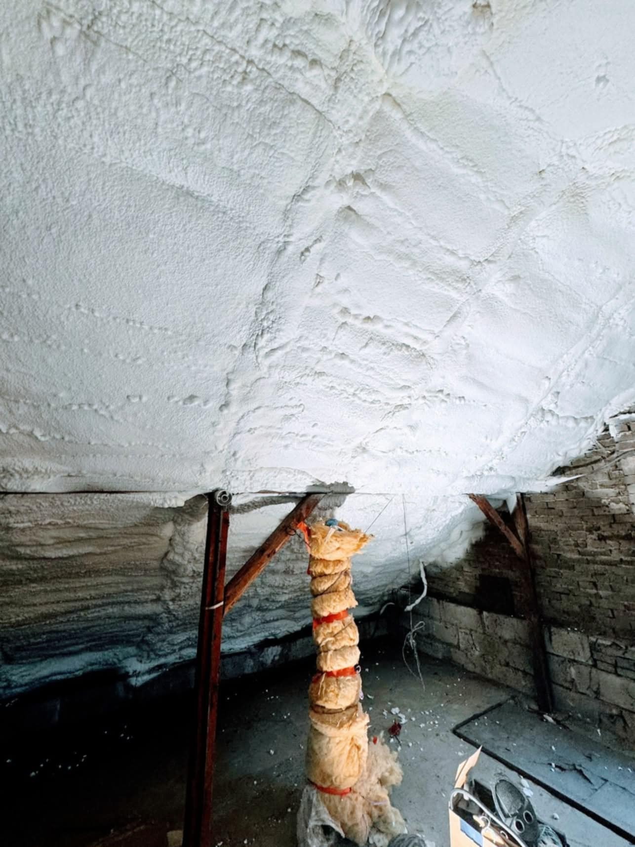 Poddasze po izolacji pianą PUR, widoczne drewniane elementy konstrukcyjne i resztki materiałów izolacyjnych. Nieregularna, biała powierzchnia izolacji.