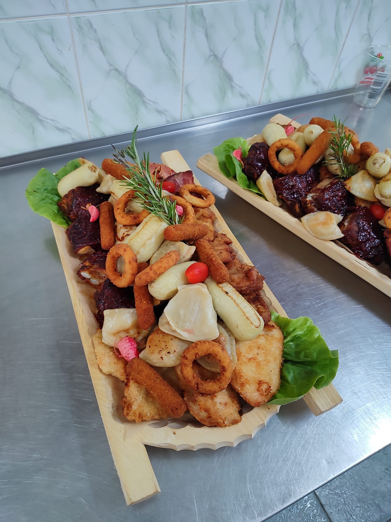 Dwa drewniane półmiski z różnorodnymi przekąskami: smażone krążki cebulowe, mięso w sosie BBQ, pieczone ziemniaki, smażone ryby, dekorowane sałatą i rozmarynem, ustawione na metalowym blacie.