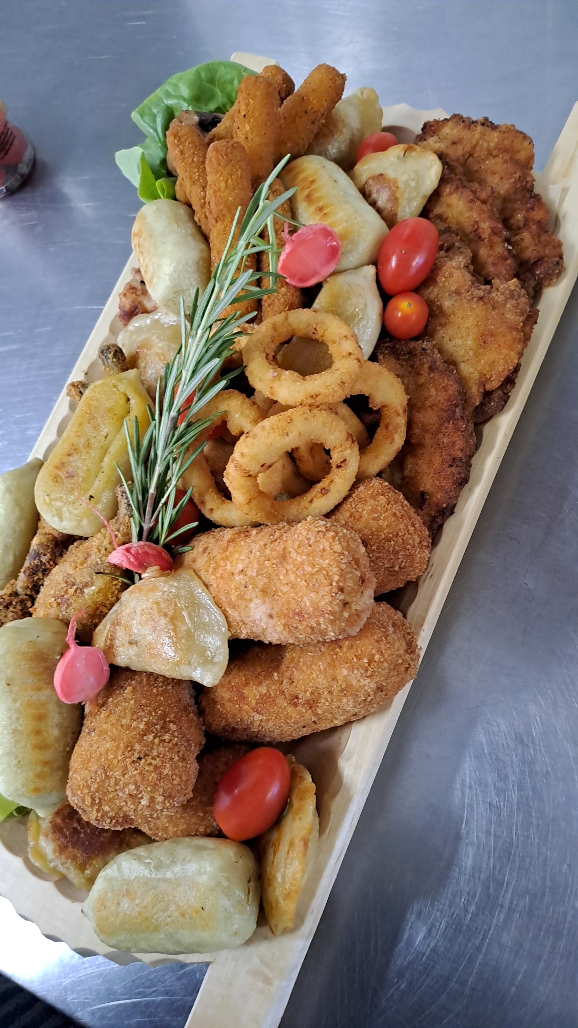 Deska z różnorodnymi przekąskami: krokiety, smażone pierogi, krążki cebulowe, kotleciki, pomidorki cherry i gałązka rozmarynu, ułożone na drewnianej desce na metalowym blacie.