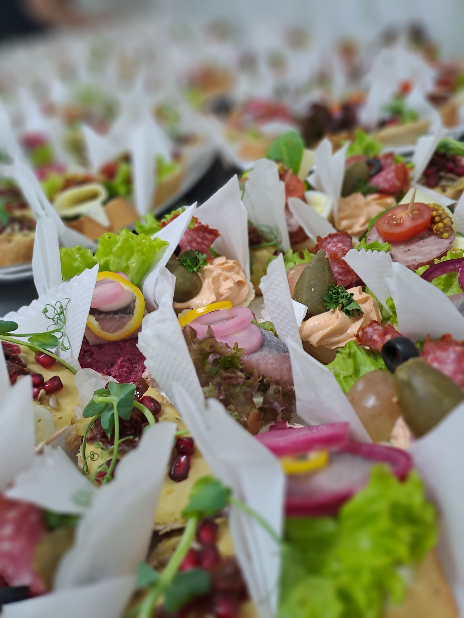 Różnorodne kanapki bankietowe, każdy kęs starannie zapakowany w papierową foremkę, prezentowane z bliska w płytkiej głębi ostrości, ukazujące bogactwo składników: od świeżych sałat, przez wędliny...