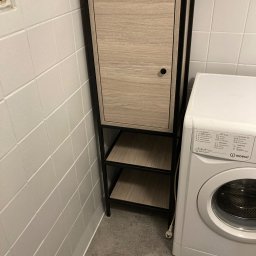 Sebastian Drewniak - Szafka łazienkowa z drzwiczkami i otwartymi półkami, o konstrukcji metalowej w kolorze czarnym i frontach w odcieniu jasnego drewna, stojąca obok pralki w pomieszczeniu wyłożonym białymi kafelkami.