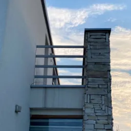 Nowoczesny balkon z prostą, metalową barierką i kamiennym filarem na tle błękitnego nieba z chmurami.