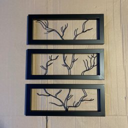 Sebastian Drewniak - Trzy czarne, prostokątne ramy z metalowymi, stylizowanymi drzewami w środku, ułożone pionowo na kartonowym tle.