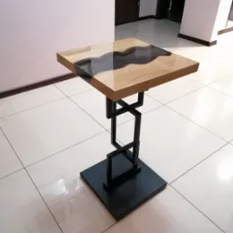 Stolik kawowy z blatem z drewna i żywicy epoksydowej, na metalowej podstawie o geometrycznym kształcie, ustawiony na jasnych płytkach podłogowych.