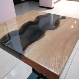 Sebastian Drewniak - Kwadratowy stolik kawowy z blatem z jasnego drewna dębowego i wtopioną, nieregularną żywicą epoksydową w kolorze antracytowym, osadzony na metalowej, czarnej podstawie w kształcie litery X.