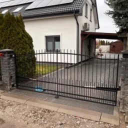 Czarna brama wjazdowa z pionowymi prętami, zamontowana na ceglanych słupkach, na tle domu z panelami słonecznymi na dachu.