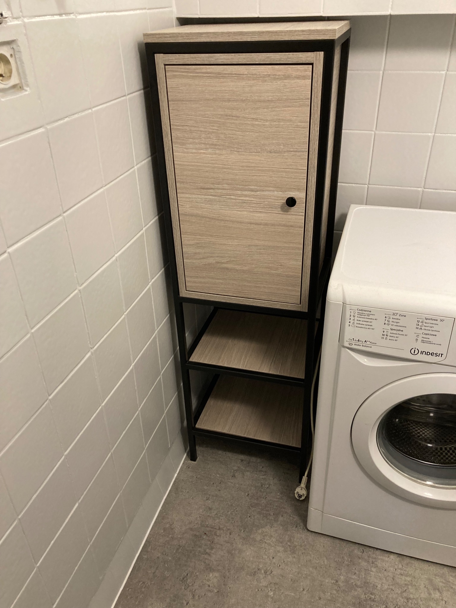 Szafka łazienkowa z drzwiczkami i otwartymi półkami, o konstrukcji metalowej w kolorze czarnym i frontach w odcieniu jasnego drewna, stojąca obok pralki w pomieszczeniu wyłożonym białymi kafelkami.