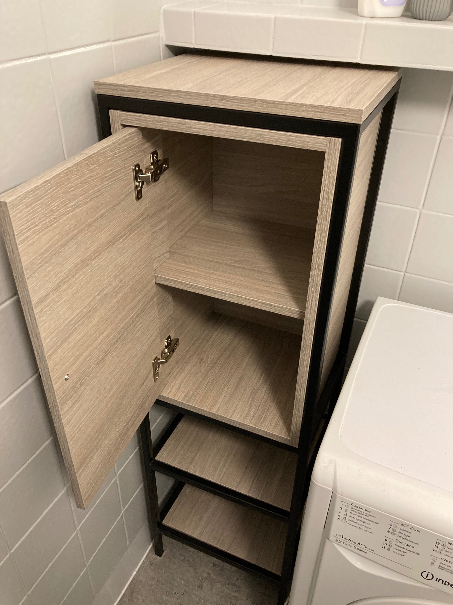 Półka łazienkowa z drewnopodobną okleiną i czarną metalową ramą, zamontowana w łazience w Tulcach obok pralki, z otwartymi drzwiczkami ukazującymi wewnętrzne półki.