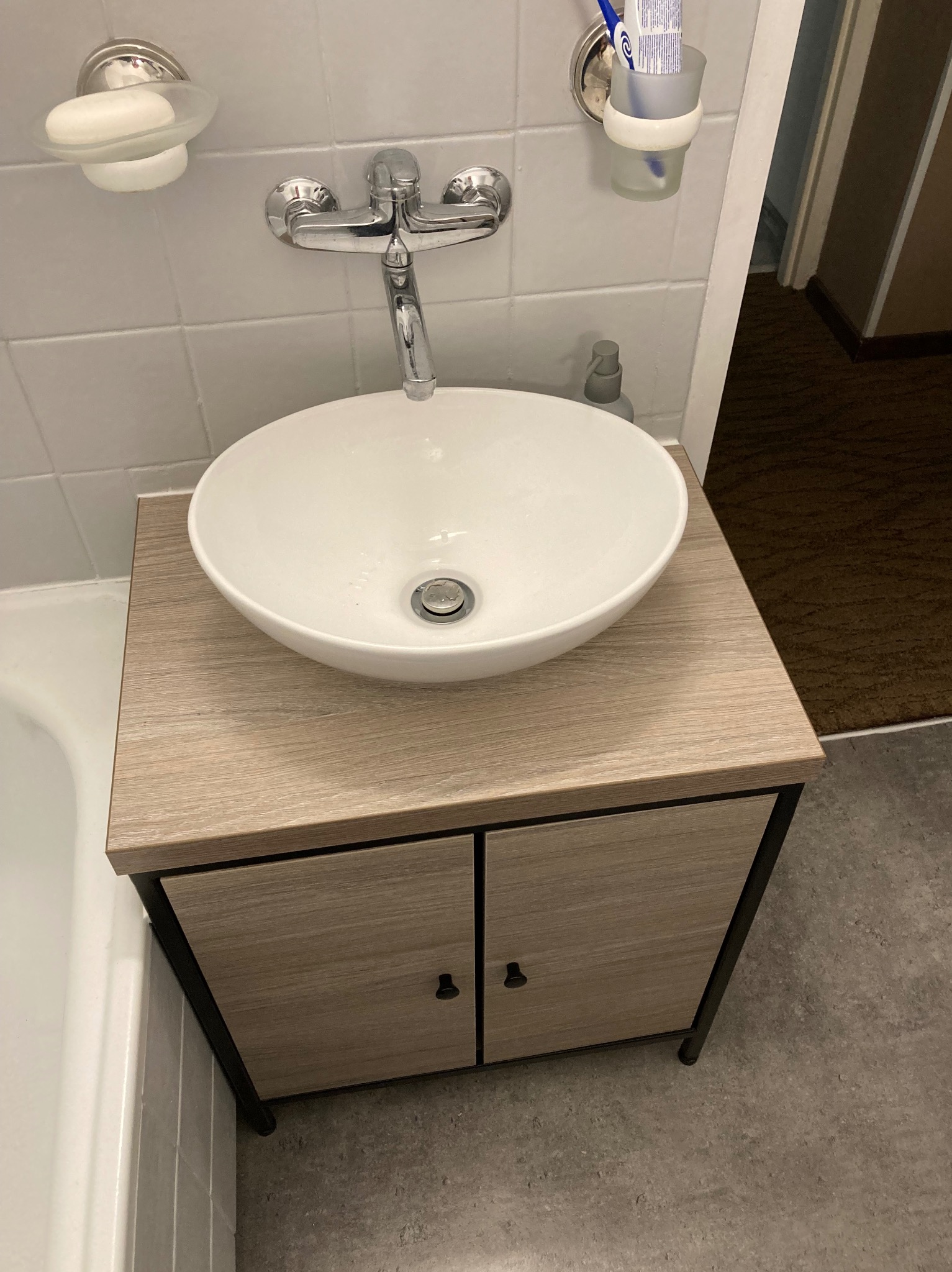 Biała umywalka nablatowa w kształcie misy, zamontowana na szafce z drewnopodobnym blatem i czarnymi nogami, bateria chromowana, mydelniczka i uchwyt na szczoteczki do zębów na ścianie z białych...