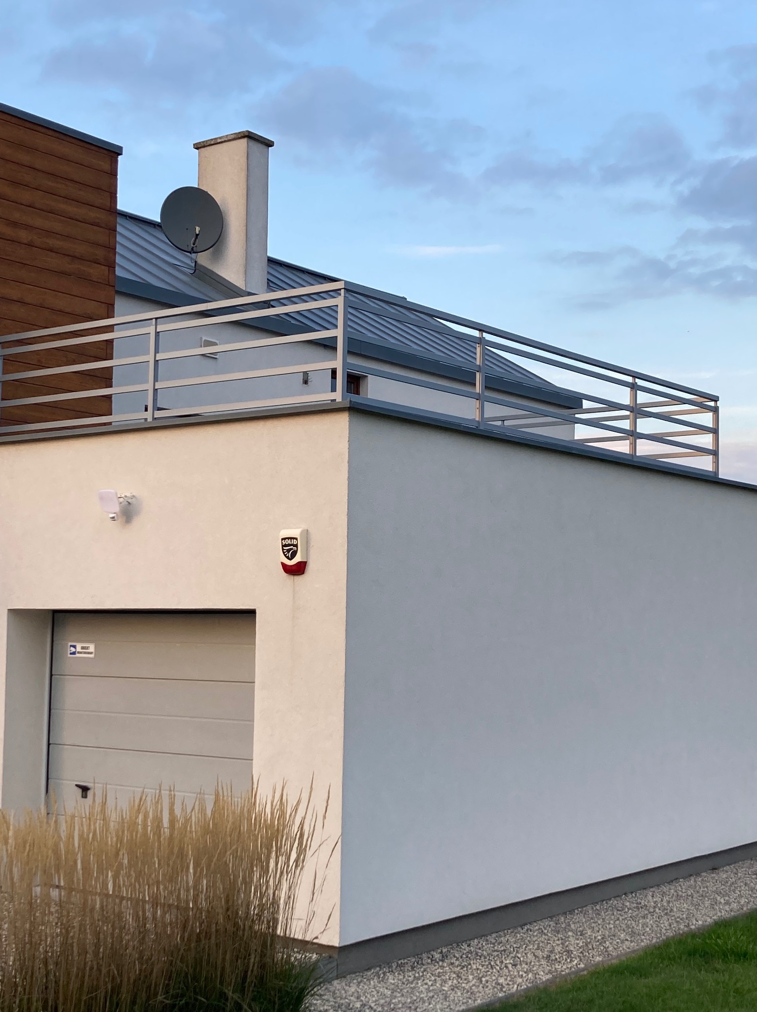 Nowoczesny budynek z płaskim dachem, metalową balustradą na dachu, bramą garażową, alarmem i lampą na elewacji, otoczony ozdobną trawą.